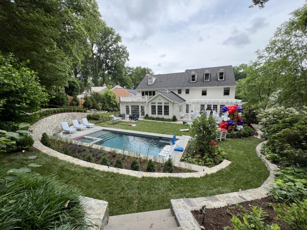 A large backyard with a private, in-ground pool, a trimmed lawn, and paved walkways. A large, two-story house sits in the background.